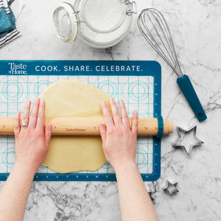 pastry mat on counter with person rolling out dough and whisk and cookie cutters to the side