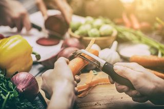 A hand holds one carrot while peeling carrot with vegetable peeler. The background features a purple onion, yellow bell pepper, and bowl of other green vegetables.