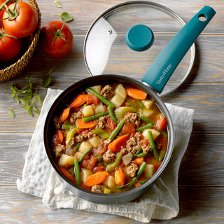 sauce pan on table with beef stew sitting next to tomatoes with lid set aside. 