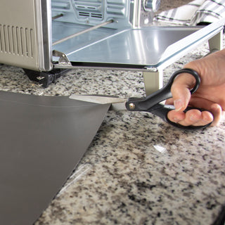 Trimming the toaster oven liner with household scissors on granite countertop next to the toaster oven.