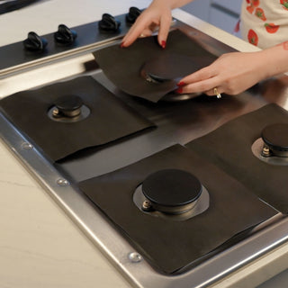 A person's hands are shown placing black gas stovetop liners on a stainless steel gas stovetop. Three of the black liners are already laid out on the stovetop burners, and the person is in the process of covering the fourth burner.