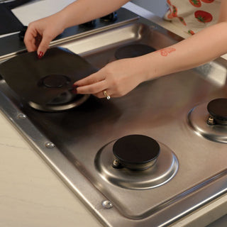 A person's hands are shown placing a black stove top liner onto a stainless steel gas stovetop.