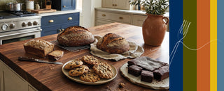 A styled kitchen scene with freshly baked bread loaves, chocolate chip cookies, and brownies arranged on a wooden countertop. A terracotta vase with greenery sits nearby, and a vertical multicolor stripe graphic with a white fork outline appears along the right edge.
