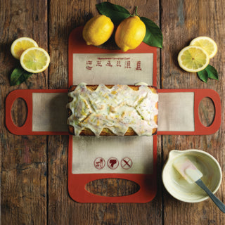 terracotta silicone baking sling laying flat on wooden table with iced lemon quick bread and accessories