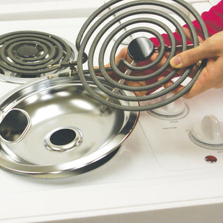 A person's hand holding a coil burner element next to a drip bowl insert on a white electric stovetop.