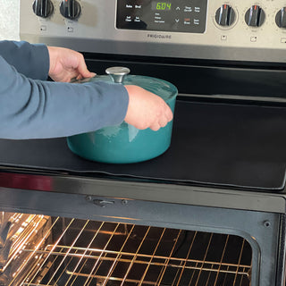 person placing a cast iron pot on the protective mat on top of smooth top range to demonstrate how the mat protects the fragile smooth top stove