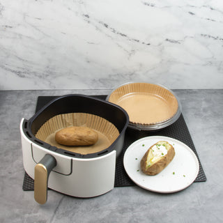 air fryer basket with baked potato in air fryer liner sitting next to baked potato with sour cream and chives and package of air fryer liners sitting on black counter mat on gray counter