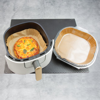 air fryer basket with cooked pizza next to package of square air fryer liners on black counter mat and gray counter