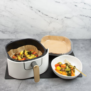 air fryer basket with cooked vegetables next to package of square air fryer liners and bowl of vegetables on black counter mat and gray counter