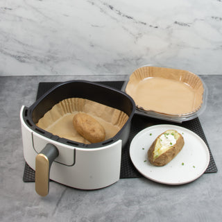 air fryer basket with baked potato next to package of square air fryer liners and plate with baked potato and sour cream on white plate on black counter mat and gray counter