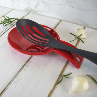 Black slotted spoon on a red spoon rest with garlic and rosemary on a white wooden surface