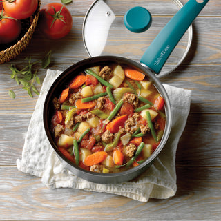 Frying pan with vegetable and meat stew on a wooden surface, surrounded by tomatoes and herbs.