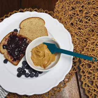 Taste of Home Mini Spatula in Sea Green on a plate with toast spread with peanut butter and jelly, surrounded by blueberries on a woven mat.