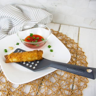 Egg roll on a taste of home vented turner with a small bowl of sauce on a white plate, on a textured surface.